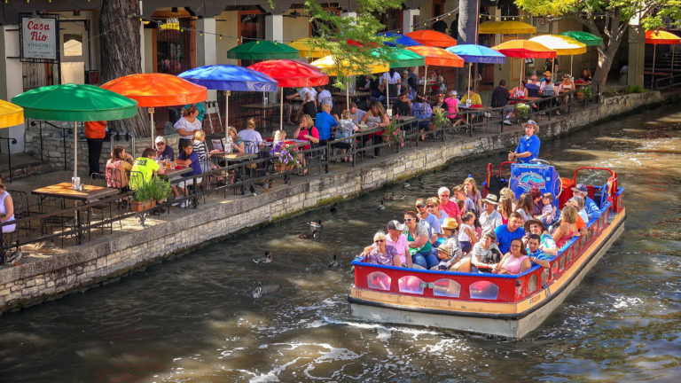 River boat on San Antonio River Walk in front of restaurant with colorful umbrellas.